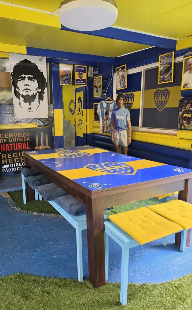 Mesa azul y amarilla con bancos, camisetas y recuerdos de Boca Juniors al fondo